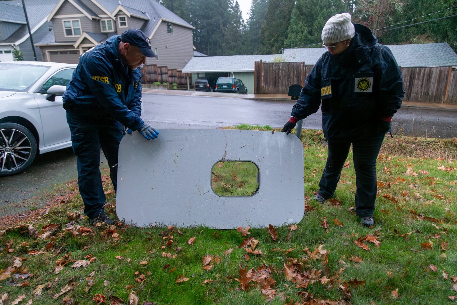 Two NTSB investigators examining the recovered door plug panel, laid on the grass at the Oregon property where it was found, its window cut-out visible.