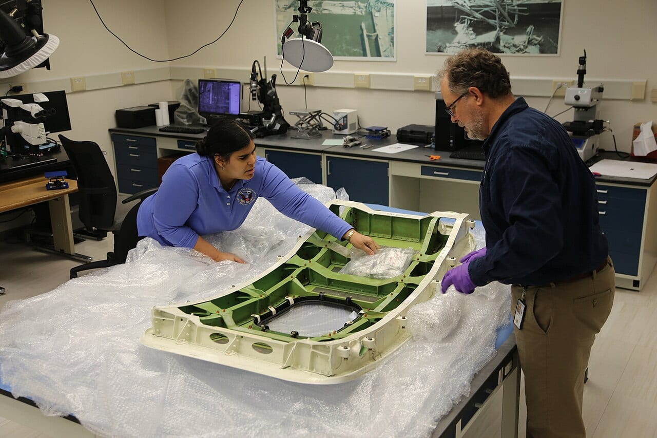 NTSB aerospace engineer and materials chief technical advisor unpacking the door plug in the NTSB Materials Laboratory in Washington, D.C., its bolt-hole fittings visible along the frame.