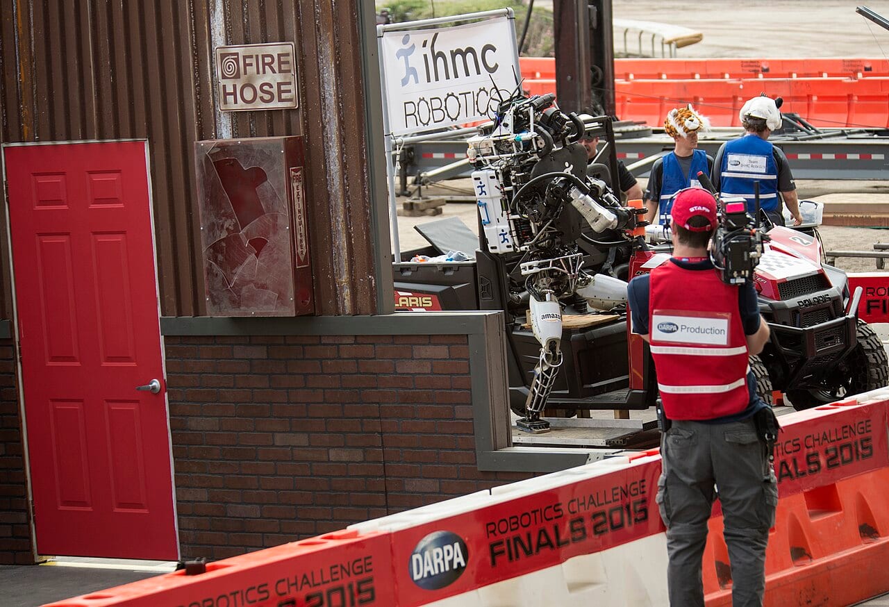 A bipedal humanoid robot climbing out of a vehicle during the 2015 DARPA Robotics Challenge, watched by its research team.