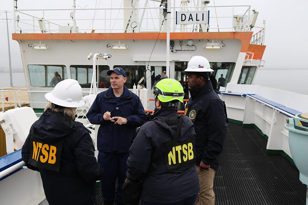 NTSB and Coast Guard investigators standing on the deck of the Dali, its name visible on the superstructure behind them, during the on-board investigation.