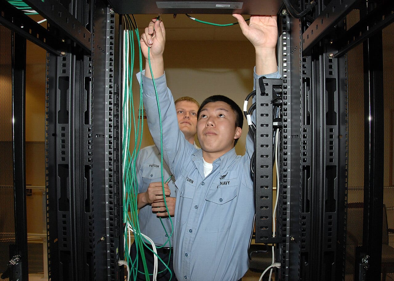 A U.S. Navy information systems technician reconfiguring network cabling while a colleague observes, on a cyber defense operations network.
